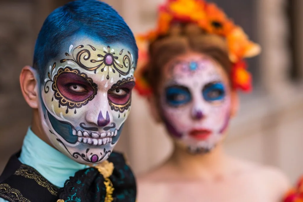 Two people wearing elaborate Día de los Muertos sugar skull face paint with intricate floral designs, colourful decorative patterns, and traditional Mexican Day of the Dead makeup celebrating cultural heritage.