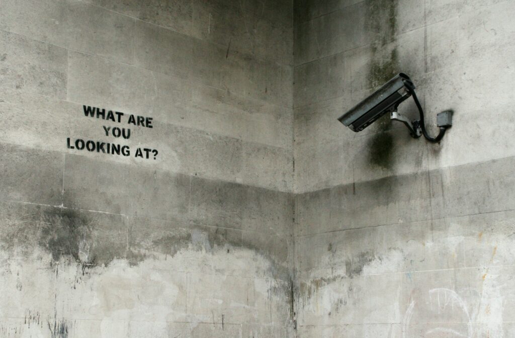 Concret walls. A corner. A CCTV camera looks at a sign that says "What are you looking at?"