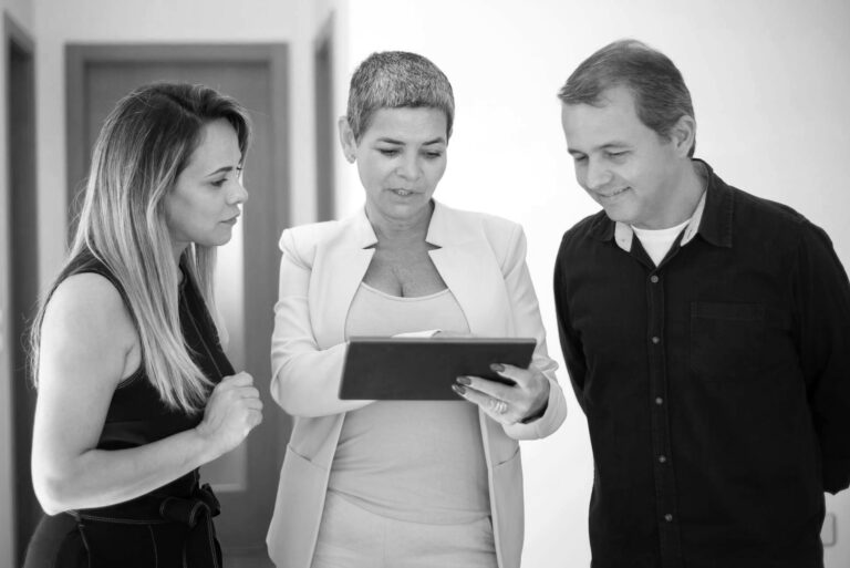 Two women and a man standing in a room. The woman in the middle is holding a tablet they are all looking at.