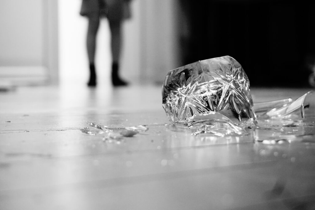 A smashed glass on the floor, in the foreground, with a pair of legs standing in the background.