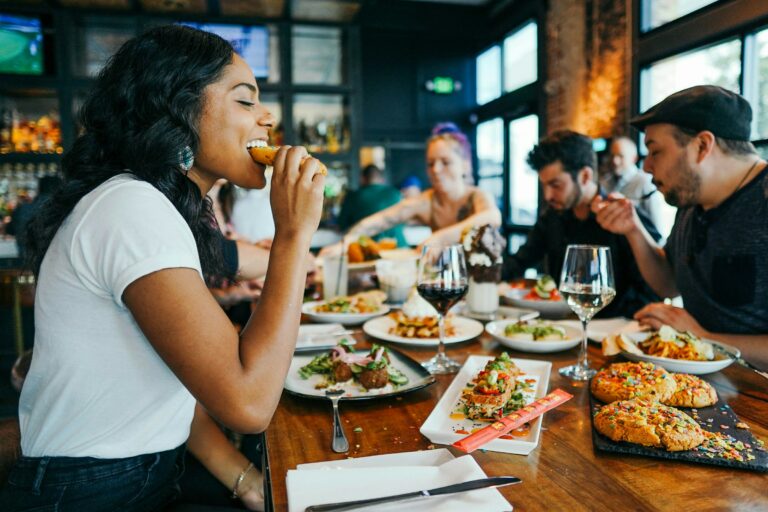 Four people around a table in a restaurant. The table has various dishes on it. A lady in the foreground in a white shirt is biting into food.