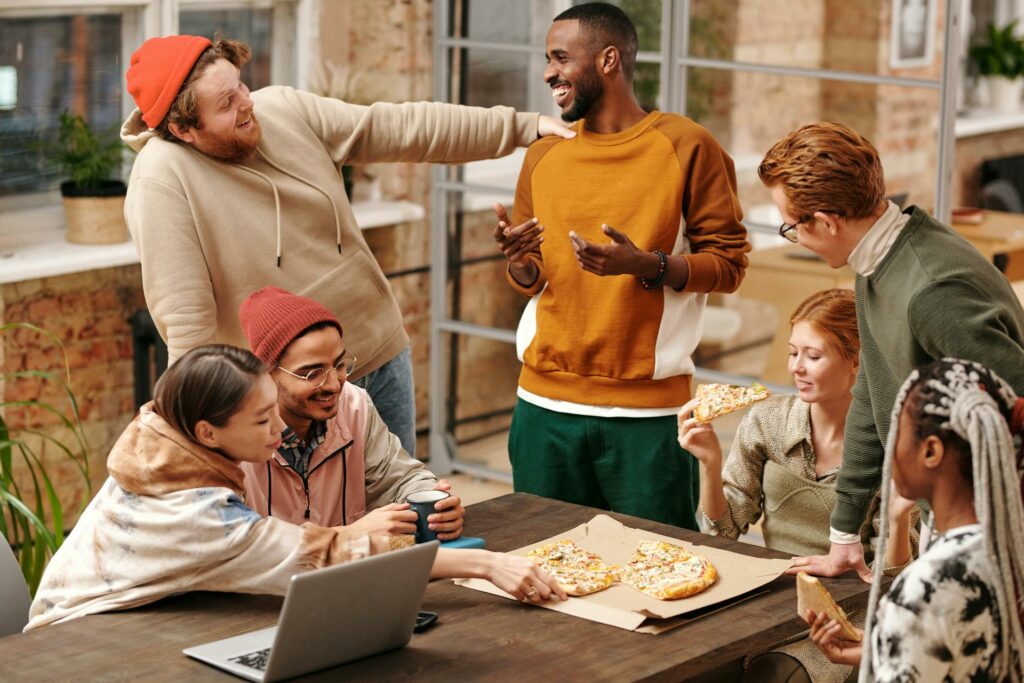 A group of people sits and stands around a table, chatting and eating pizza.