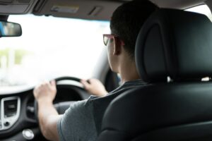 Man driving a car in the UK, wearing glasses and a gray t-shirt, viewed from the back seat, with a bright sky visible through the windscreen