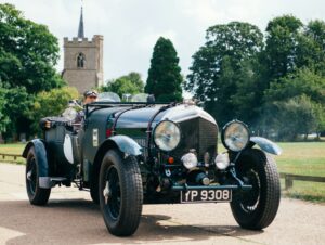 A vintage car, with its dark green paint gleaming, drives down a quiet road. A church spire rises in the background, and the driver, wearing goggles and a cap, has a joyful expression.