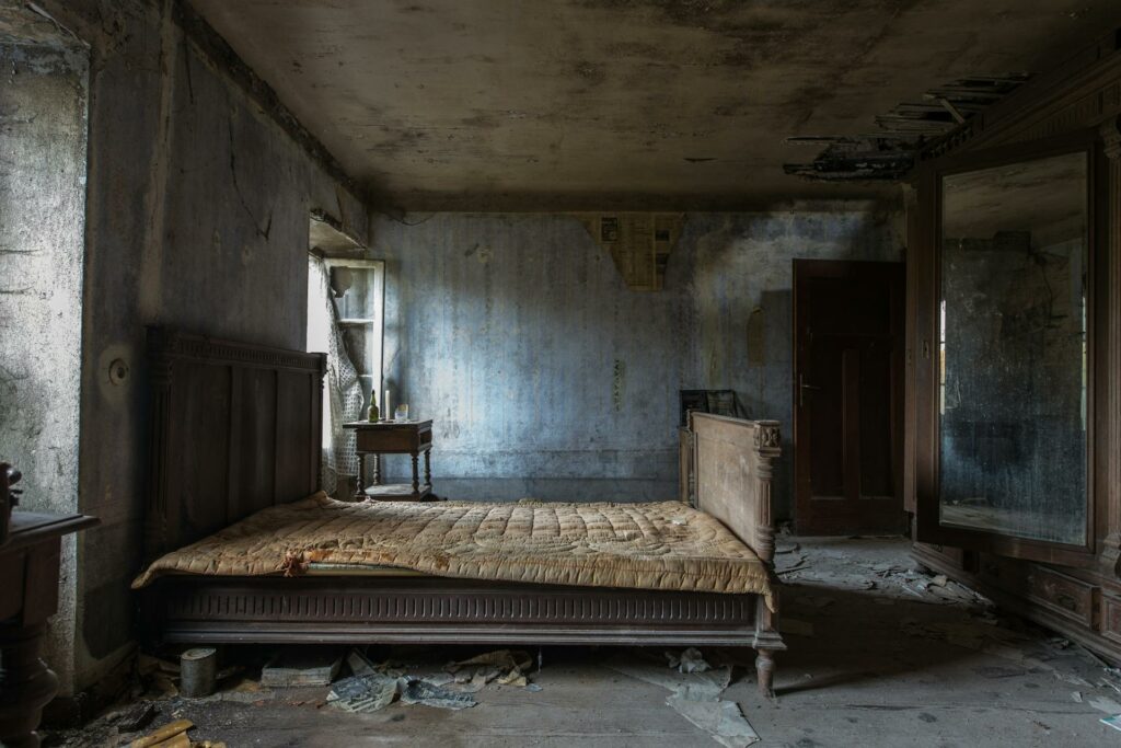 A dilapidated bedroom in an abandoned house. A large wooden bed with a tattered quilt dominates the room. Peeling wallpaper, a cracked ceiling, and debris scattered across the floor convey a sense of decay and neglect.