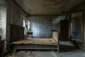 A dilapidated bedroom in an abandoned house. A large wooden bed with a tattered quilt dominates the room. Peeling wallpaper, a cracked ceiling, and debris scattered across the floor convey a sense of decay and neglect.