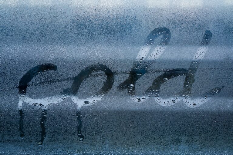 Condensation streaks down a window pane. The word "cold" is written in the condensation, highlighting the temperature difference causing the water droplets.