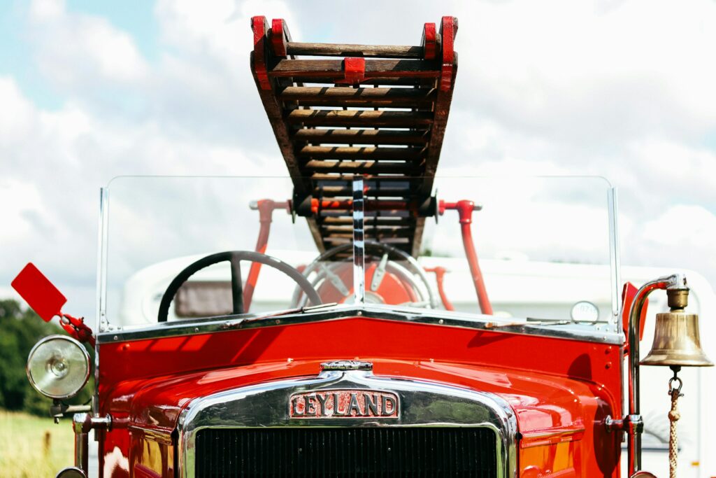 A close-up view of a vintage red Leyland fire engine. The gleaming chrome grill and hood ornament are prominent, with the manufacturer's name "LEYLAND" prominently displayed. A wooden ladder is extended upwards above the truck, and a brass bell hangs on the side.