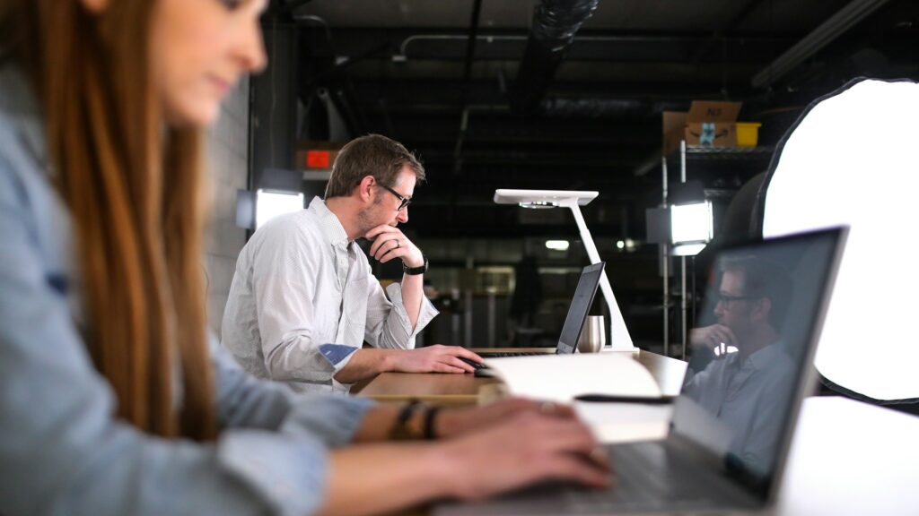 A man and a woman working in a modern office, both focused on their laptops. The man in the background types thoughtfully, while the woman in the foreground is blurred, concentrating on her screen. The space is well-lit with a minimalist design.