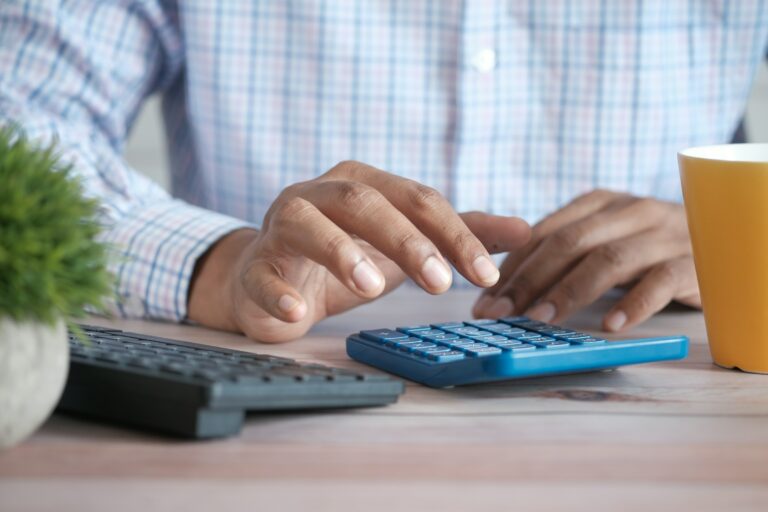 A person uses a blue calculator on a light wood desk. A computer keyboard, green plant, and yellow cup are also visible on the desk.