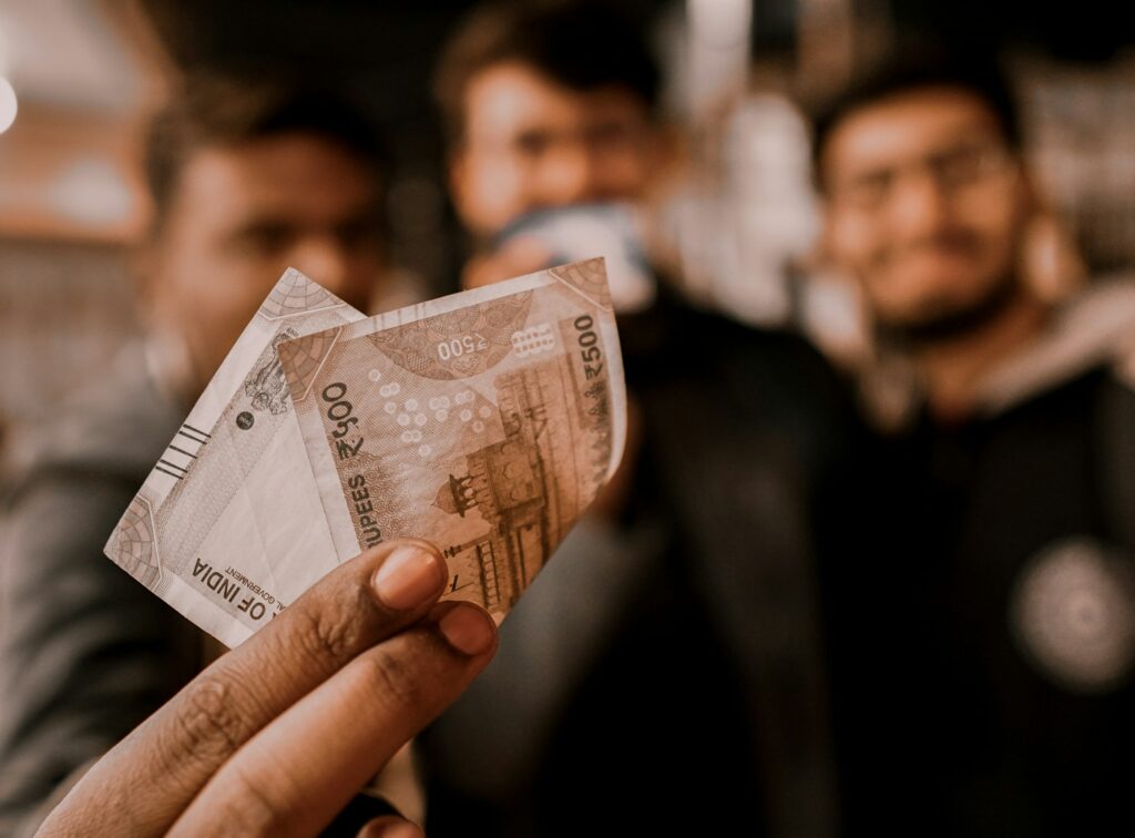 The image shows a hand holding a 500 Indian Rupee banknote in the foreground, with blurred people visible in the background.