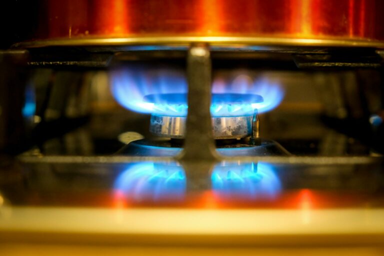 A close-up view of a lit gas burner on a stovetop, with bright blue flames and a red pot visible on the hob.