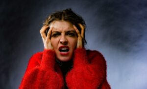 A person in a red fuzzy coat showing extreme frustration, with hands pressed to their temples and mouth open in a distressed expression against a dark background.