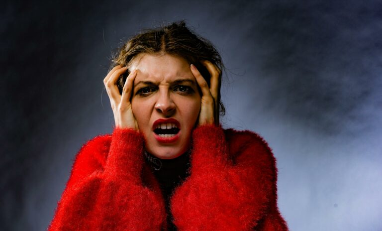 A person in a red fuzzy coat showing extreme frustration, with hands pressed to their temples and mouth open in a distressed expression against a dark background.