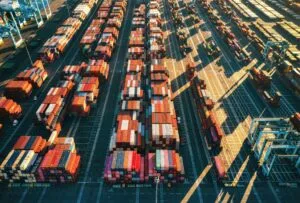 Aerial view of a shipping container terminal with rows of colorful containers neatly arranged across the port. Gantry cranes are visible at the edges, with afternoon sunlight casting long shadows across the facility.