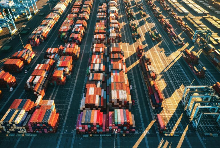Aerial view of a shipping container terminal with rows of colorful containers neatly arranged across the port. Gantry cranes are visible at the edges, with afternoon sunlight casting long shadows across the facility.