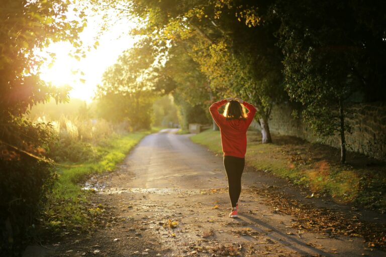 The image shows a person in a red hoodie walking along a path through a wooded area. The path is surrounded by lush greenery and the sunlight is filtering through the trees, creating a warm, golden atmosphere. The person appears to be enjoying a peaceful stroll through the natural setting.