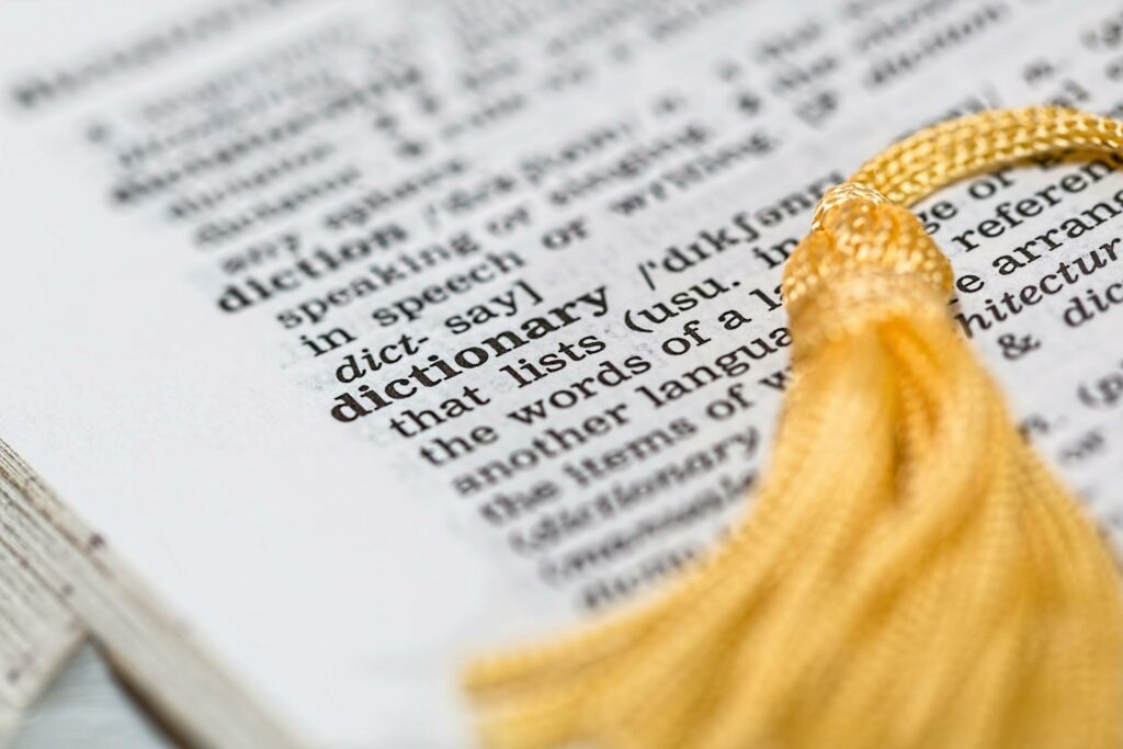 A close-up of a dictionary page, with the focus on the definition of the word "dictionary". The page is partially obscured by a yellow tassel, which appears to be from a bookmark.