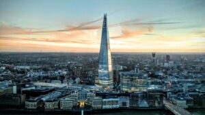 Aerial view of South London at sunset, dominated by the Shard skyscraper in the centre. The glass pyramid-shaped building reflects golden evening light against a pink and blue sky. The surrounding cityscape shows a mix of modern and historic buildings along the Thames River.