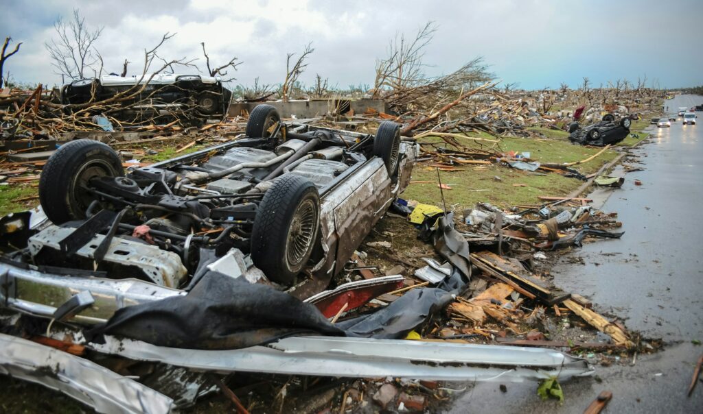 Devastating aftermath of a tornado showing extensive climate impact destruction: overturned and damaged vehicles, widespread debris, splintered trees, and demolished structures stretching across a residential area. A car lies upside down in the foreground amidst scattered wreckage and storm damage on a rain-soaked street.