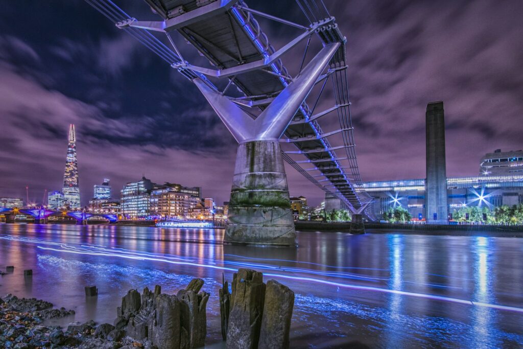 A dramatic night-time view of London's Millennium Bridge and the River Thames. The bridge is illuminated in blue light, with The Shard skyscraper visible in the background against a purple-hued sky. The Thames water reflects the city lights, creating streaks of blue light on its surface. In the foreground, old wooden posts protrude from the riverside, and the Tate Modern's chimney is visible on the right. The cityscape is lit up with office buildings and the bridge's modern architecture contrasts with the historical elements along the river.