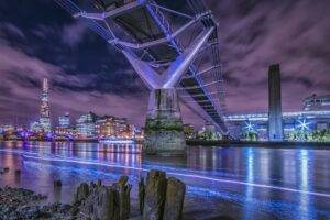 A dramatic night-time view of London's Millennium Bridge and the River Thames. The bridge is illuminated in blue light, with The Shard skyscraper visible in the background against a purple-hued sky. The Thames water reflects the city lights, creating streaks of blue light on its surface. In the foreground, old wooden posts protrude from the riverside, and the Tate Modern's chimney is visible on the right. The cityscape is lit up with office buildings and the bridge's modern architecture contrasts with the historical elements along the river.