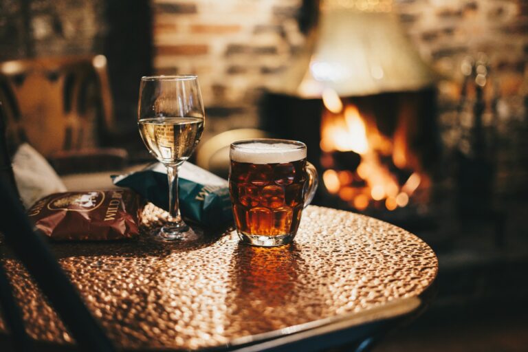 A cozy pub setting with a textured copper table holding a glass of white wine and a pint of dark beer or ale. A packet of crisps is visible on the table. In the background, a warm fireplace burns, creating a golden glow throughout the space. The scene has a comfortable, intimate atmosphere typical of a traditional British pub, with soft lighting and rustic elements like brick walls partially visible in the blurred background.