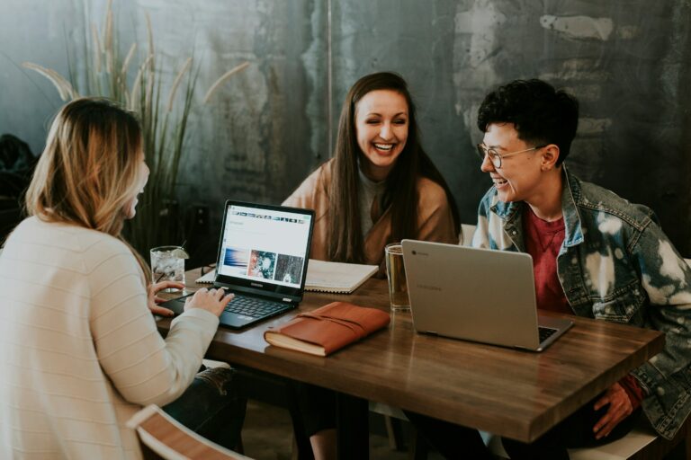 Three people sitting at a wooden table in a casual setting, engaged in a work meeting. They have laptops open and appear to be collaborating cheerfully. One person wears a cream sweater, another a tan coat, and the third a denim jacket with red shirt. The setting has industrial-style décor with textured walls and decorative plants.