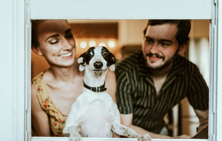 A small black and white dog (likely a Jack Russell terrier) with its paws on a windowsill, looking at the camera. Two smiling people stand behind the dog - one wearing a floral top and the other in a striped shirt. The scene is framed by a white window frame with warm lighting in the background.