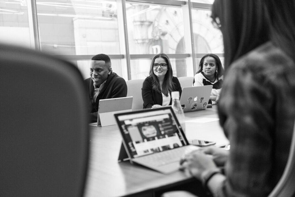 Black and white image of a business meeting with three professionals seated at a table with laptops and tablets. They're facing a person whose back is to the camera. The setting is a bright office with large windows, suggesting a collaborative work environment.