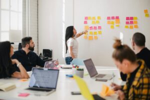 A woman points at sticky notes on a wall during a relocation strategy workshop, while colleagues sit at a table with laptops in a bright, modern office space.