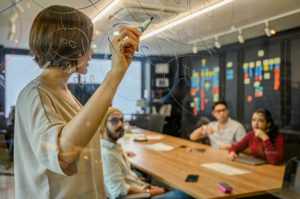 A person in a beige top writes on a transparent glass wall or board during what appears to be a business meeting. They are viewed from behind while drawing or writing diagrams, with three colleagues seated at a wooden table in the background. The room has modern office lighting and colourful post-it notes visible on a dark wall in the distance.