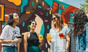 Four women holding craft beers, laughing together in front of a vibrant, multicoloured street art mural. Dressed casually, they appear to be enjoying a social moment in an urban setting.