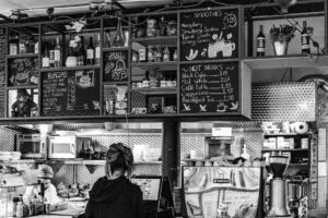 A black and white photo of a small London café interior showing a staff member at the counter. Chalkboard menus display the prices for various items including burgers, smoothies, and hot drinks. The kitchen area and storage shelves with bottles and supplies are visible in the background.