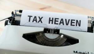 Close-up image of a white typewriter with a sheet of paper inserted that has "TAX HEAVEN" typed in large, bold black letters. The typewriter appears to be vintage style with its metal typebars visible beneath the paper and traditional keyboard keys visible at the bottom of the frame. The typewriter sits on a light wooden surface.