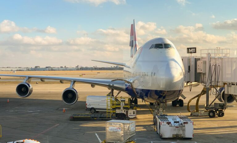 A British Airways Boeing 747 aircraft parked at gate M11 at Chicago O'Hare International Airport Terminal 1. The plane is connected to a jetway with various ground service vehicles and cargo containers surrounding it. The image shows a clear day with blue skies and scattered clouds, with the airport tarmac visible.