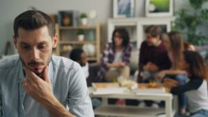 A pensive man in a light blue shirt sits in the foreground, hand on chin, looking downcast. In the background, a blurred group of people are seated around a table in a modern, minimalist office or home setting with wooden shelves, plants, and framed artwork visible.