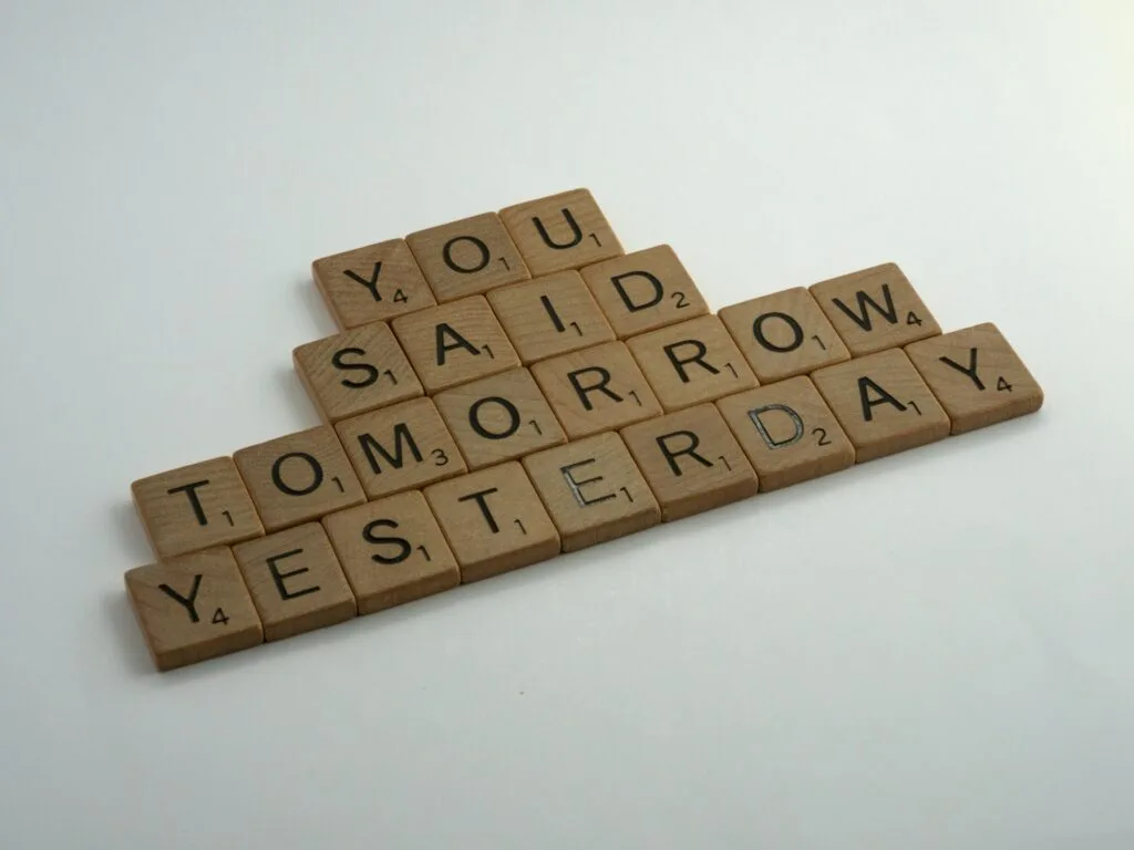 Wooden Scrabble tiles arranged to spell out "YOU SAID TOMORROW YESTERDAY" against a light background, conveying a message about procrastination and delays.