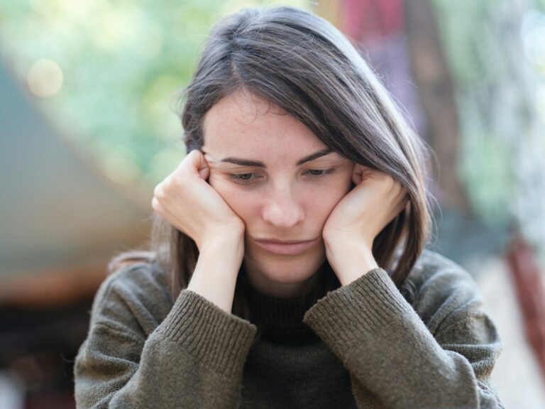 The image shows a person with long dark hair looking downcast with their face resting in their hands. They appear to be in a contemplative or distressed state, wearing a grey jumper in what seems to be an indoor setting with a blurred background. The posture and facial expression suggest feelings of sadness, worry, or exhaustion.