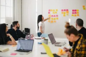 Woman pointing at colorful sticky notes arranged on a white wall during a meeting, while colleagues with laptops observe her presentation in a bright, modern office space.