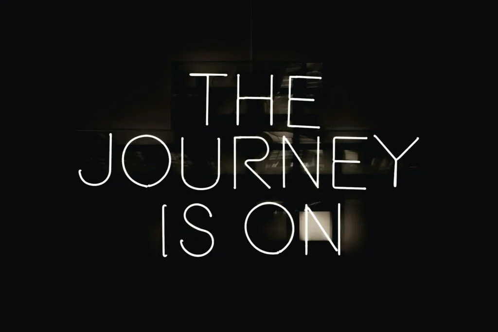A black background with white neon text that reads "THE JOURNEY IS ON" displayed across three lines. The text has a glowing effect typical of neon signage, creating a stark contrast against the dark background.
