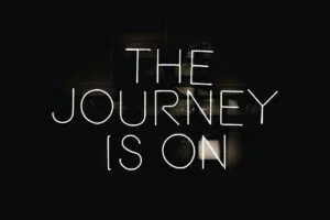 A black background with white neon text that reads "THE JOURNEY IS ON" displayed across three lines. The text has a glowing effect typical of neon signage, creating a stark contrast against the dark background.