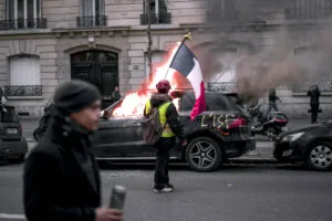 A person with a backpack and French flag stands near a car with flames coming from the interior. Smoke is visible, and other people are in the background, hinting at a scene of civil unrest.