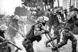 Riot police in full tactical gear advancing during what appears to be a protest. Officers wear black uniforms, helmets with face shields, and carry riot shields and weapons. The scene is smoky with urban buildings in the background. The image captures police in formation during a tense confrontation.RetryClaude can make mistakes. Please double-check responses.