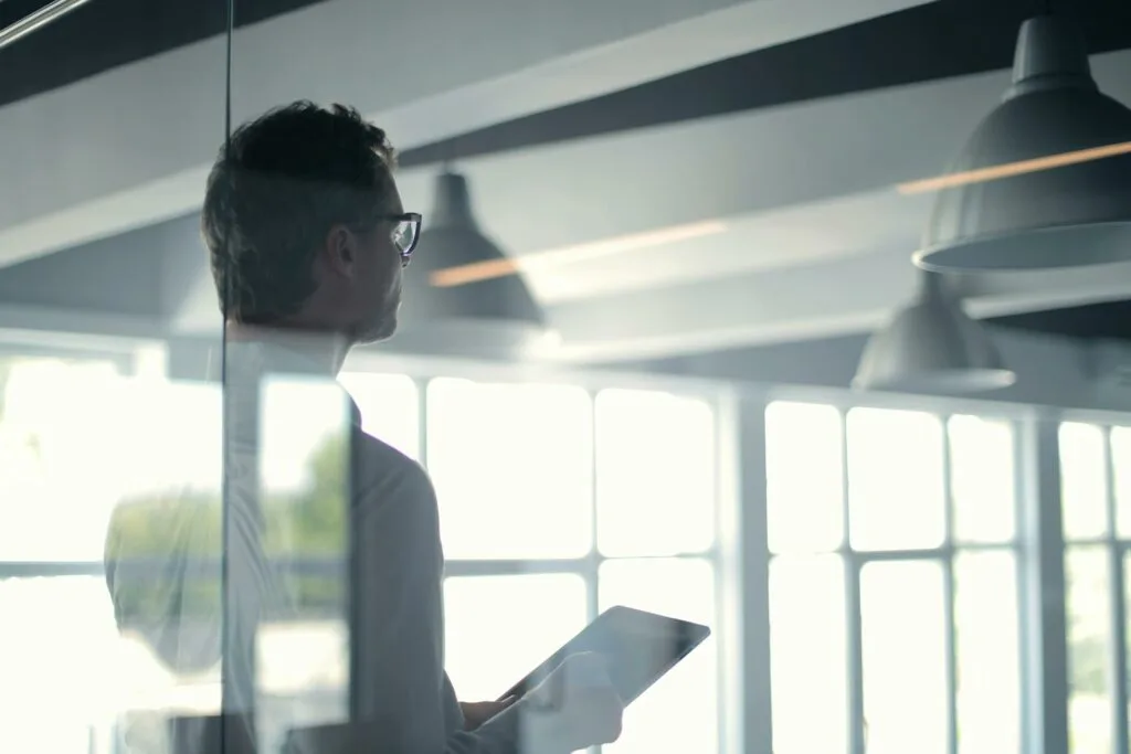 The image shows a person with glasses and an undercut hairstyle in profile, holding a tablet in a modern office space. They're looking toward large windows with natural light streaming in. Industrial pendant lights hang overhead, and the scene is captured through reflective glass, creating a bright, contemporary workplace atmosphere.