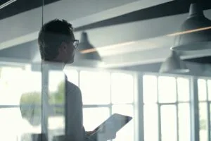 The image shows a person with glasses and an undercut hairstyle in profile, holding a tablet in a modern office space. They're looking toward large windows with natural light streaming in. Industrial pendant lights hang overhead, and the scene is captured through reflective glass, creating a bright, contemporary workplace atmosphere.