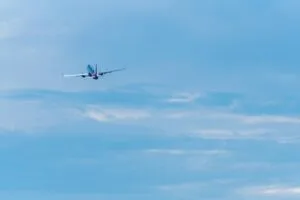 The image shows a commercial passenger aircraft in flight against a clear blue sky with some thin, wispy clouds. The plane appears to be ascending after takeoff, with its distinctive blue and white livery visible. It's captured from below, displaying the aircraft's underside as it flies against the bright daytime sky.