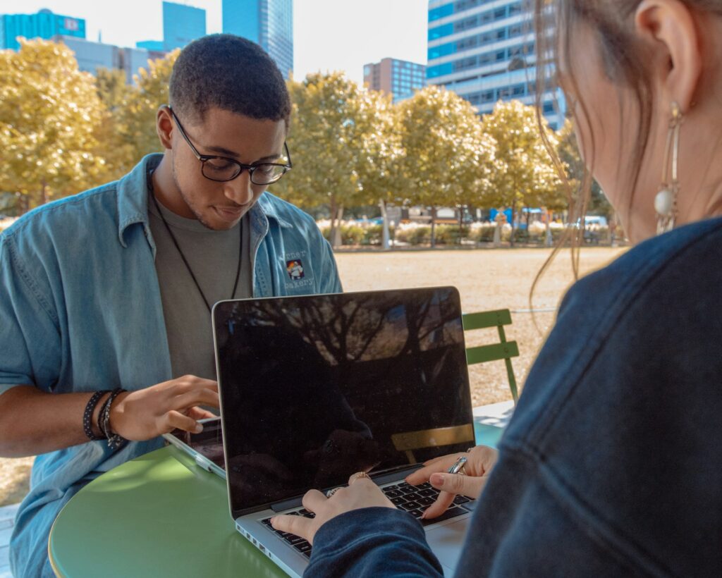 Two young adults collaborating on a laptop in an urban park. Man in blue denim shirt and glasses sits opposite a colleague with long hair. Autumn trees and city buildings visible in background.RetryClaude can make mistakes. Please double-check responses.