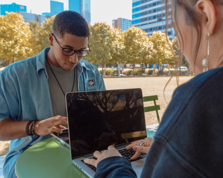 Two young adults collaborating on a laptop in an urban park. Man in blue denim shirt and glasses sits opposite a colleague with long hair. Autumn trees and city buildings visible in background.RetryClaude can make mistakes. Please double-check responses.