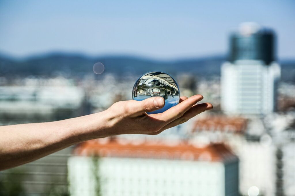 A hand holds a crystal ball reflecting an inverted cityscape against a blurred mountain backdrop.